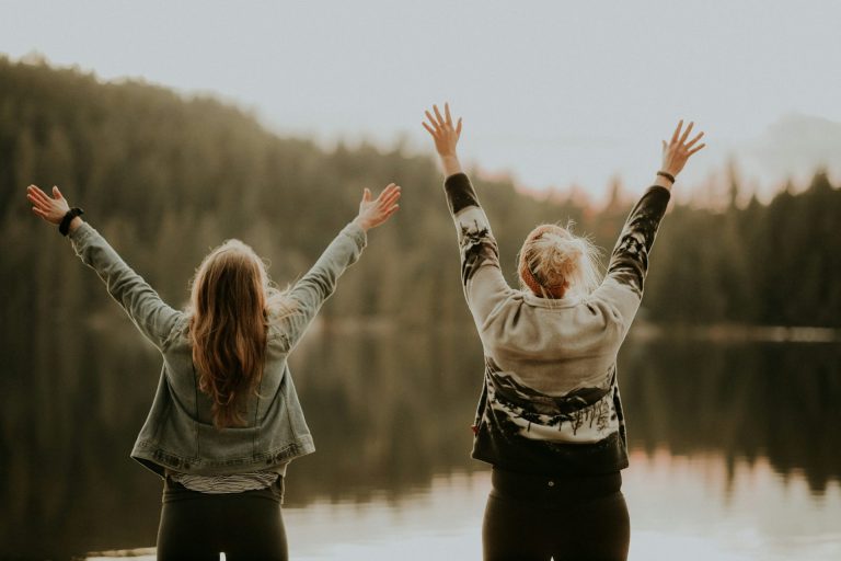 Zwei erfolgreiche Frauen stehen am Ufer eines Sees und strecken beide Hände in den Himmel.